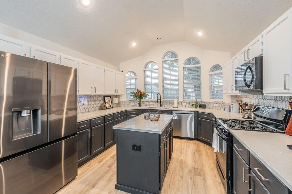 Kitchen featuring dark cabinets, black appliances, light wood-type flooring, light stone counters, and lofted ceiling