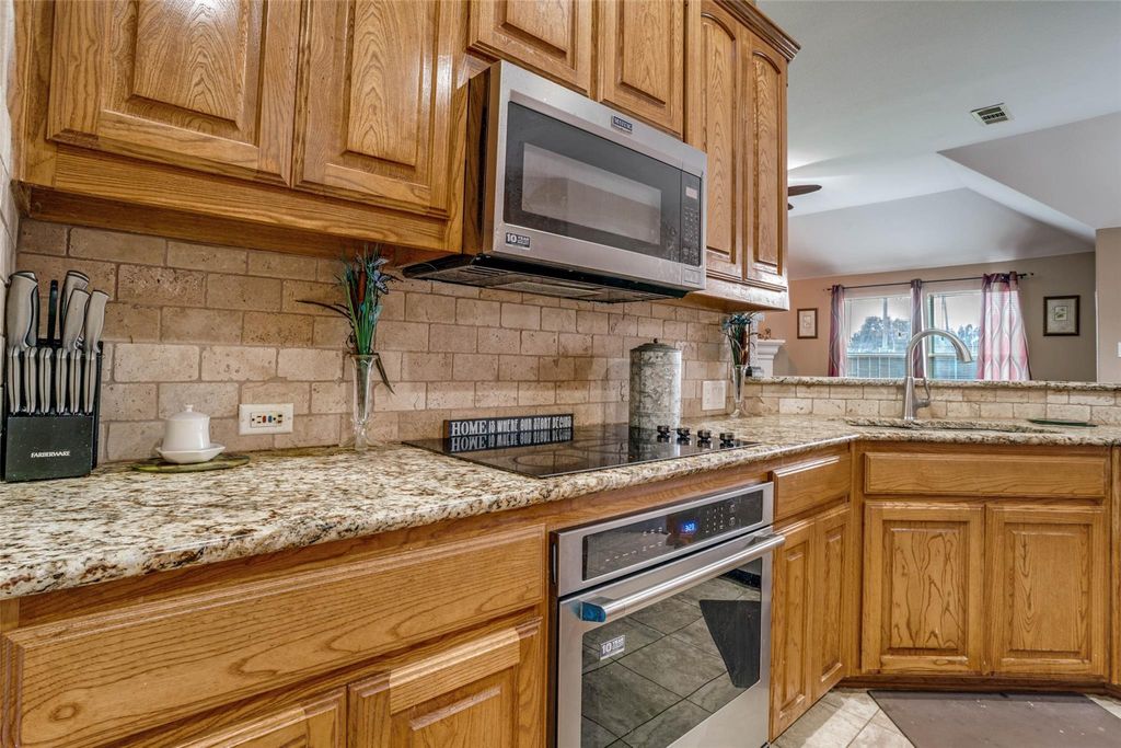 Kitchen featuring stainless steel appliances, decorative backsplash, brown cabinets, light stone counters, and light tile patterned flooring