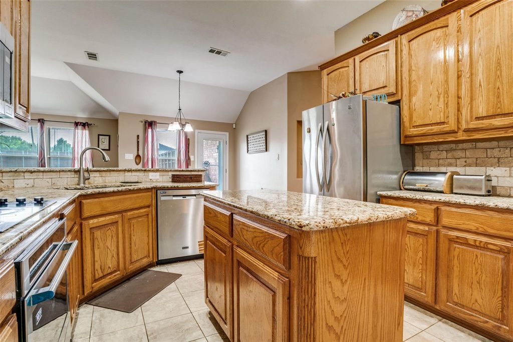 Kitchen featuring backsplash, vaulted ceiling, light stone countertops, appliances with stainless steel finishes, and brown cabinets
