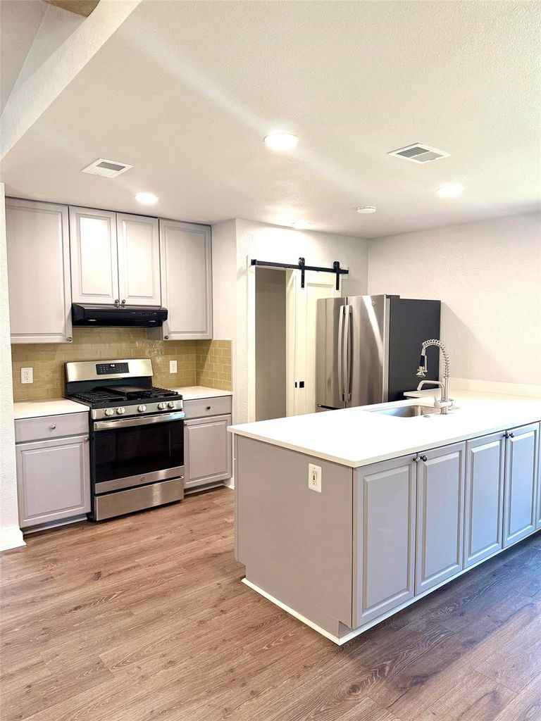 Kitchen with a barn door, backsplash, appliances with stainless steel finishes, light wood-type flooring, and an island with sink