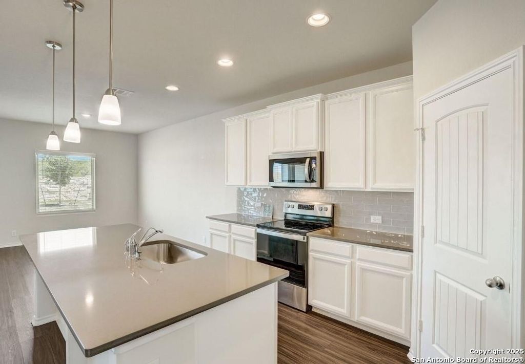 Kitchen featuring appliances with stainless steel finishes, white cabinetry, an island with sink, backsplash, and hanging light fixtures