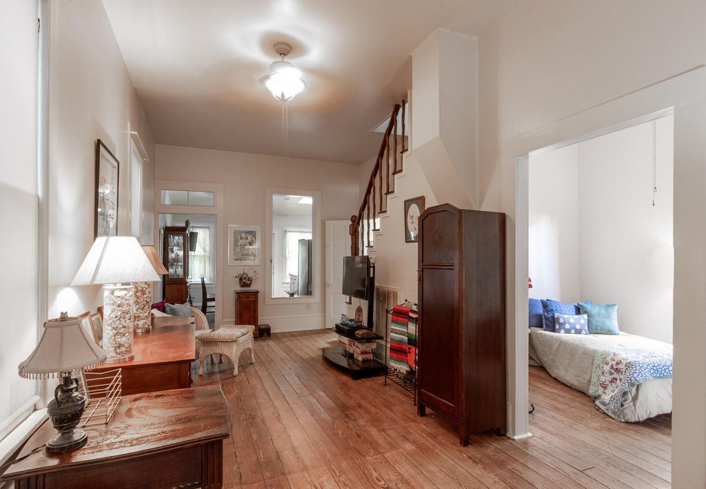 Looking from the front foyer into the living room with the second bedroom to the right. Staircase to upstairs in the right background by the downstairs bathroom door. Kitchen was added on with the doorway & transom and open window space in the background. Original wood floors and tall ceilings.