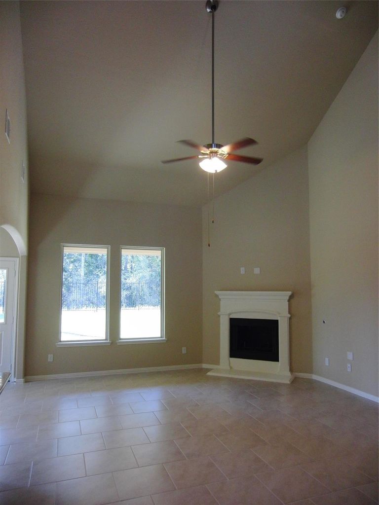 This spacious living room features high ceilings, a ceiling fan, a cozy fireplace, and large windows that provide plenty of natural light. The neutral tile flooring and walls offer a versatile backdrop for any style of decor.