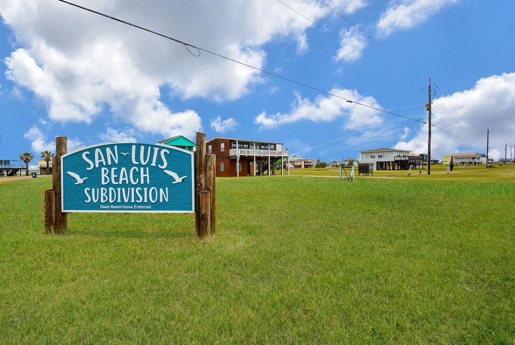 Welcome to San Luis Beach Subdivision, featuring open grassy areas with homes in the background under a bright blue sky. The sign highlights deed restrictions, indicating a well-maintained community.