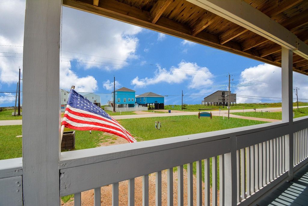 This photo showcases a view from a covered deck, featuring a neatly maintained lawn and distant houses, including a striking blue one. An American flag adds a patriotic touch, and the scene is set against a backdrop of a bright, partly cloudy sky.