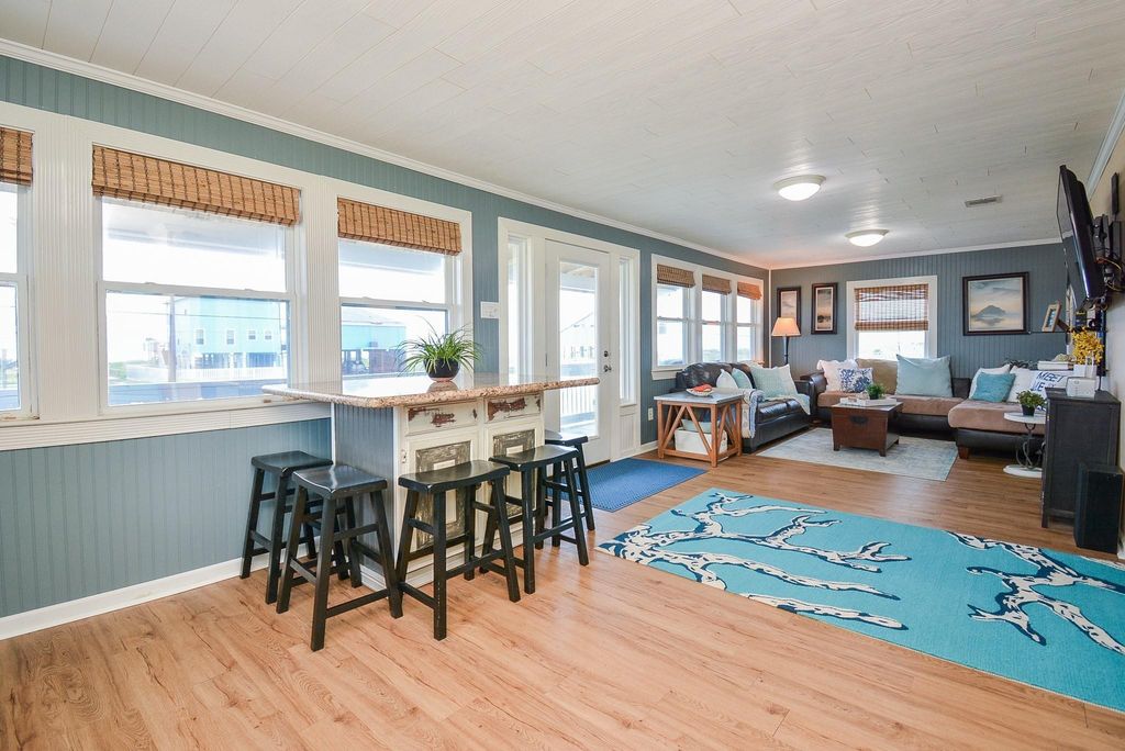 This bright, airy living space features large windows, light wood flooring, and a cozy seating area. The open layout includes a breakfast island with seating, and the room has a relaxed, coastal vibe, ideal for comfortable living and entertaining.