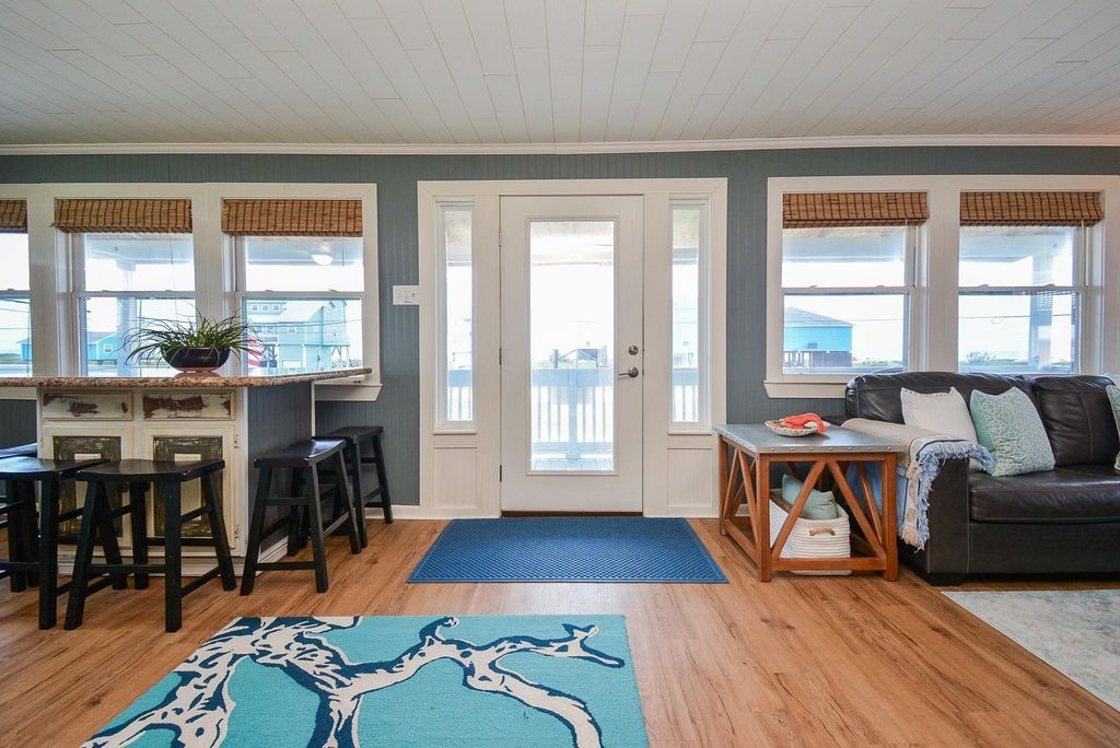 This inviting living space features large windows and a glass pane door providing abundant natural light. It includes a seating area with a sofa, a stylish area rug, and a breakfast island with stools, offering a blend of comfort and functionality for entertaining.