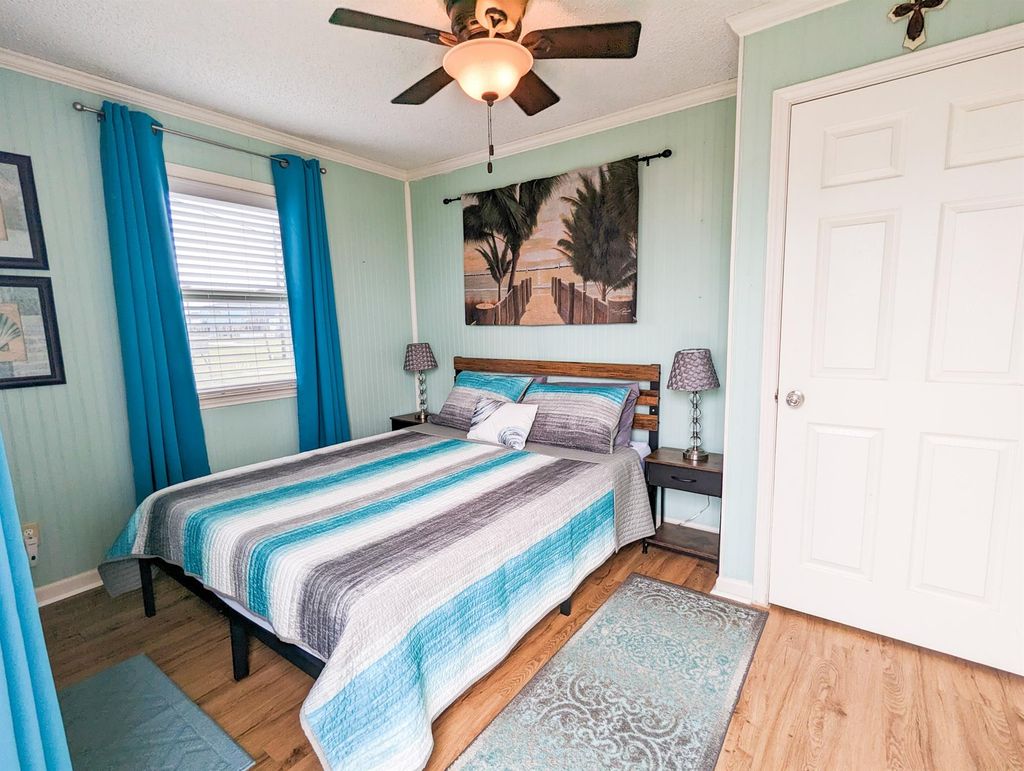 Bedroom with a beach-themed decor, featuring a double bed with a striped comforter and seating futon. There's a ceiling fan and ample natural light from a window. A beach artwork above the bed adds a relaxing touch.