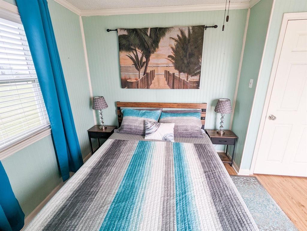 This cozy bedroom features a comfortable bed with a striped quilt, flanked by two bedside tables with lamps. The room is painted in a soothing light green, complemented by blue curtains. A beach-themed artwork adds a relaxing touch.