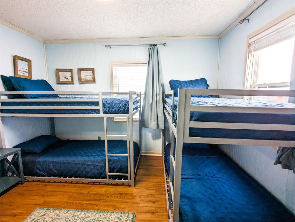This bedroom features two sets of bunk beds with blue bedding, ideal for maximizing space. The room is well-lit with natural light from two windows and has light blue walls, creating a bright and inviting atmosphere.