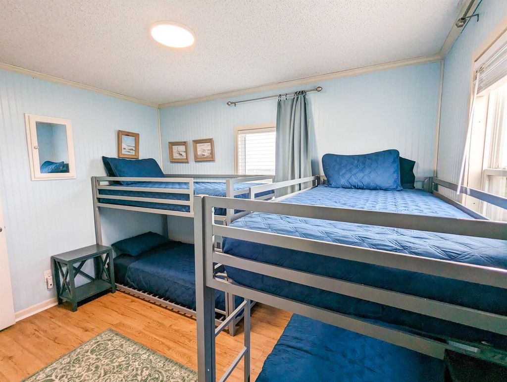 This bedroom features twin bunk beds with blue bedding, and natural light comes through two windows, complemented by a side table and decorative wall art.