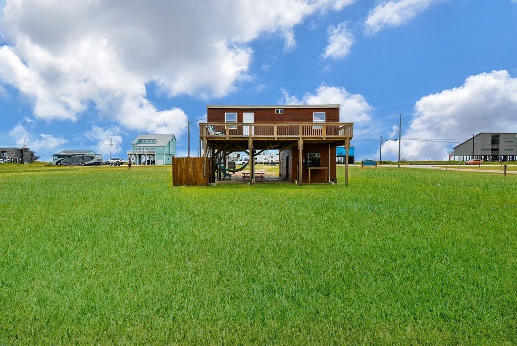 This photo shows a stilted wooden house with a spacious deck, set on a large grassy lot. The area is open with a few neighboring homes, under a bright blue sky with scattered clouds. Ideal for those seeking a serene, beach setting.