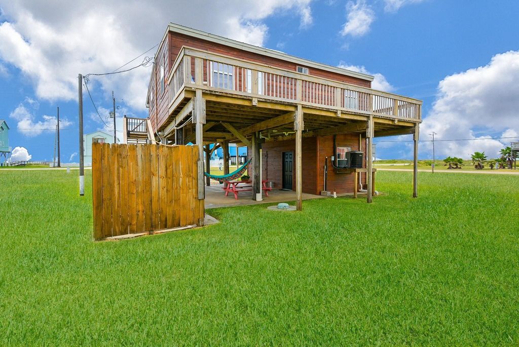 This photo shows a raised beach-style home with a spacious deck, situated on a large grassy lot. The lower level features a wooden privacy fence and a hammock and outdoor shower. The house has a coastal vibe, ideal for enjoying the outdoors.
