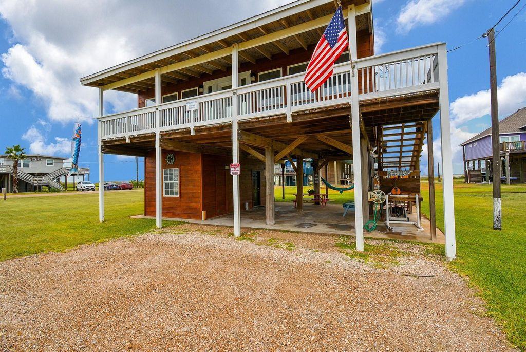 This elevated beach house features a spacious wraparound deck, an American flag, and ample covered parking beneath. It's set on a grassy lot with nearby homes, offering a coastal, relaxed vibe.