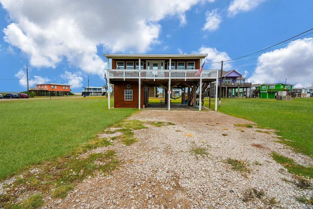 This photo shows a raised beach house with a spacious front porch, set on a grassy lot. The house has a gravel driveway and is elevated on stilts, typical for coastal areas. It's surrounded by other colorful elevated homes, with a bright, open sky above.