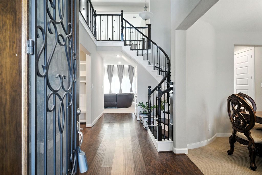 A welcoming entryway with elegant hardwood floors and a curved staircase with wrought iron railings. The open layout leads to a bright living area with large windows, and there’s a glimpse of a formal dining space on the right.