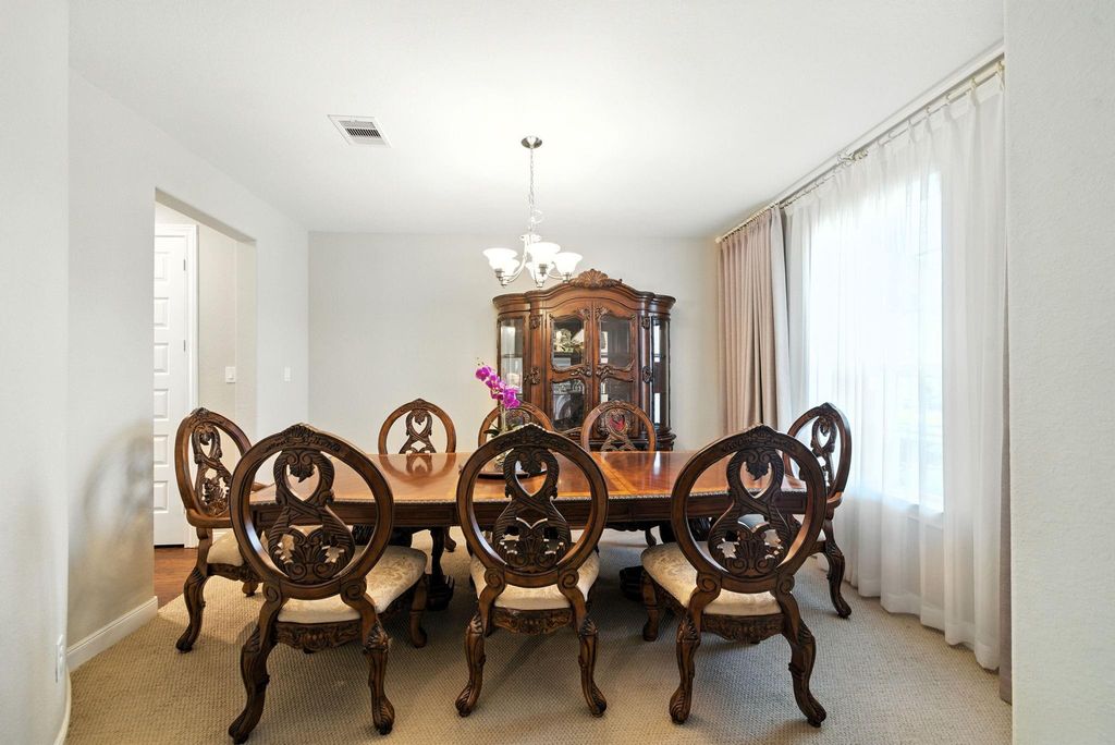Elegant dining room with a large wooden table and intricately carved chairs. Features a glass-fronted china cabinet, chandelier lighting, and a large window with sheer curtains, creating a bright and inviting space.