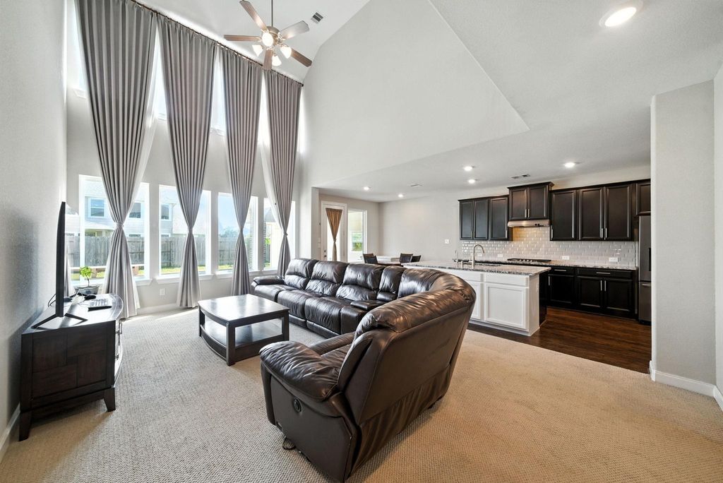 Spacious living room features high ceilings and large windows with elegant drapes, allowing plenty of natural light. It flows seamlessly into an open kitchen with dark cabinetry, a modern backsplash, and a central island, perfect for entertaining.