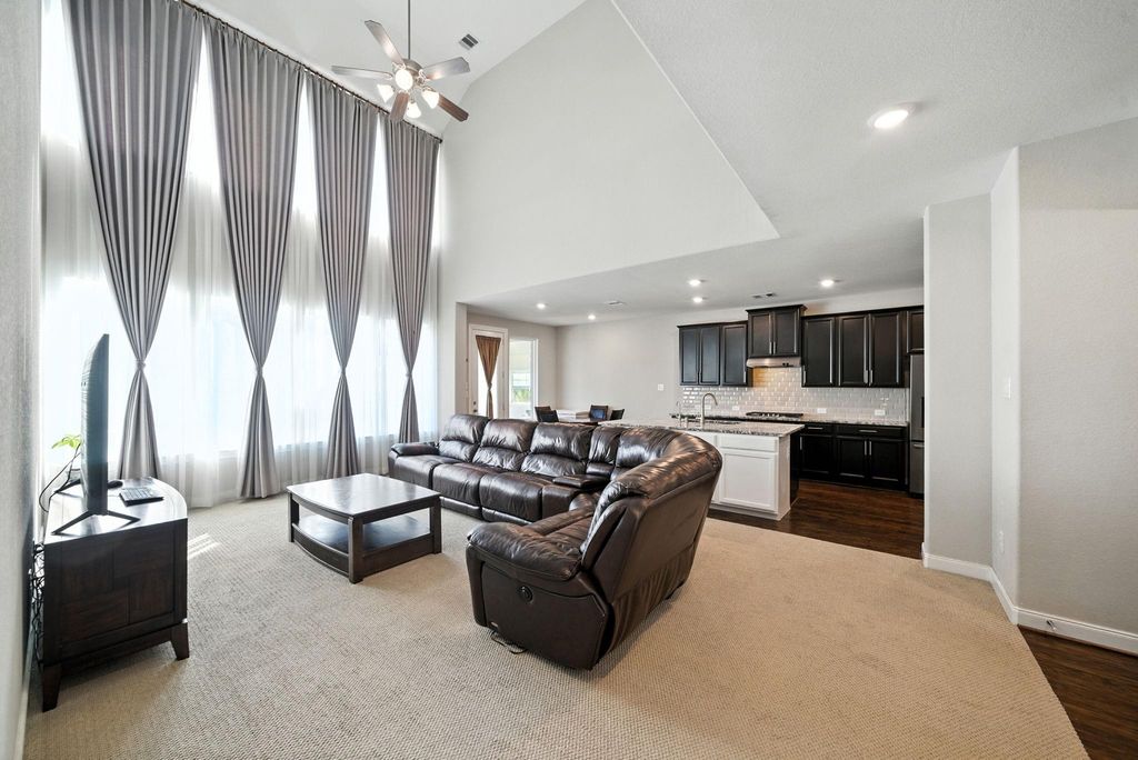 This spacious living room features high ceilings and floor-to-ceiling windows with elegant drapes, offering abundant natural light. The open layout includes a cozy seating area with a leather sectional and a sleek kitchen with dark cabinetry and modern appliances, providing a perfect setting for relaxation and entertaining.