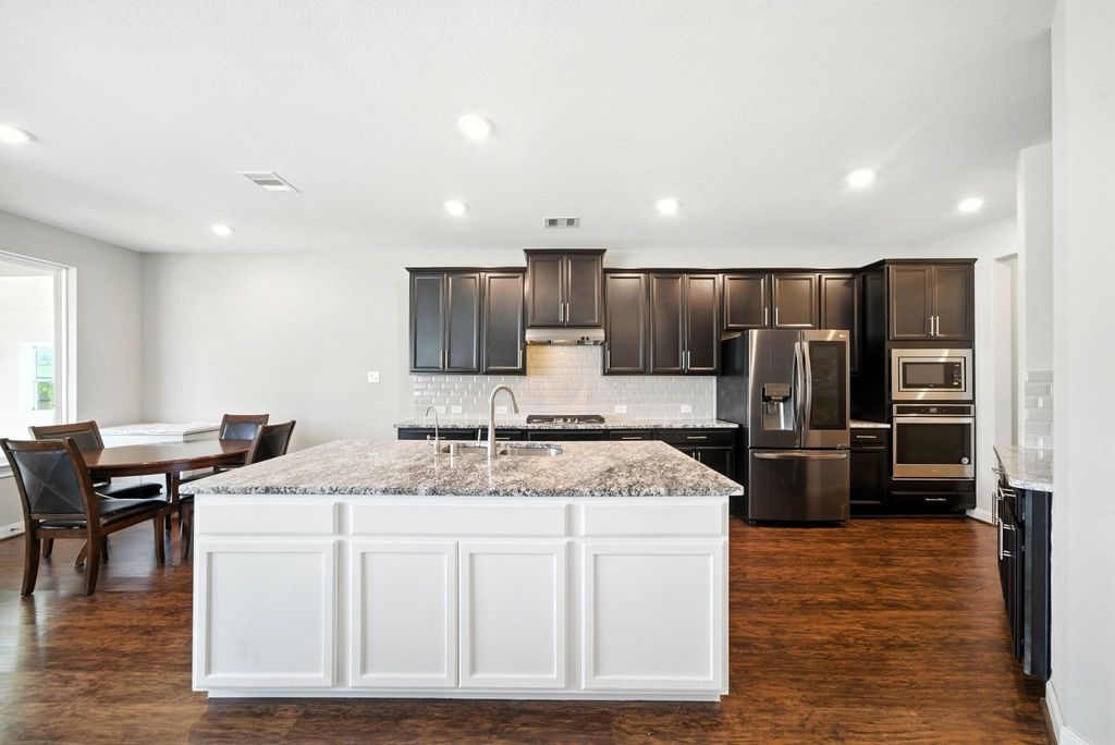 This modern kitchen features dark cabinetry, stainless steel appliances, and a spacious island with a granite countertop. The open layout connects to a dining area with wood flooring and ample natural light.