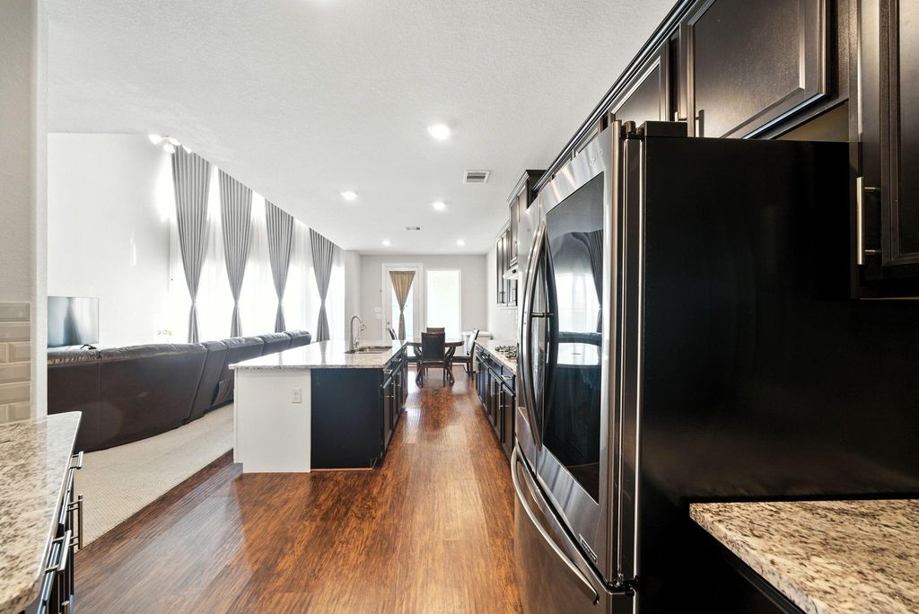 A modern, open-concept kitchen and living area. The kitchen features sleek dark cabinetry, stainless steel appliances, and granite countertops. Adjacent is a spacious living room with high ceilings and large windows, providing ample natural light. The layout flows seamlessly into a dining area, creating a welcoming and functional space.