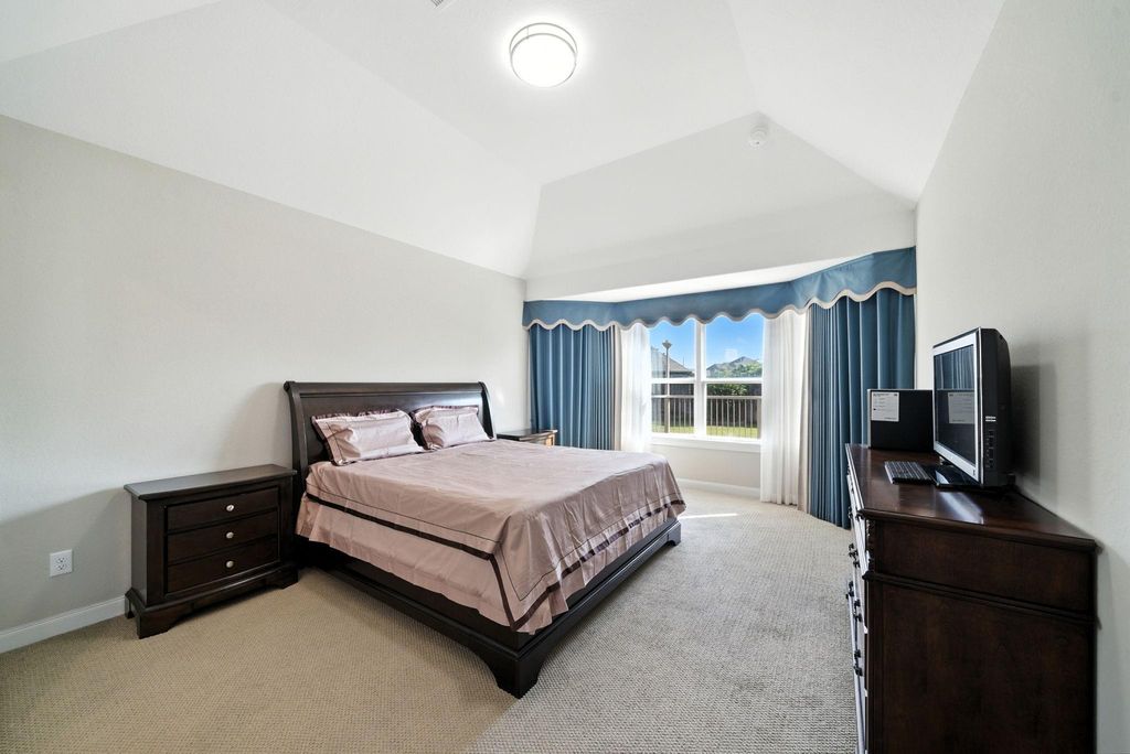 Spacious master bedroom with vaulted ceiling, featuring a large window with blue curtains allowing natural light. It includes a dark wood bed and matching furniture, creating a cozy and elegant atmosphere.