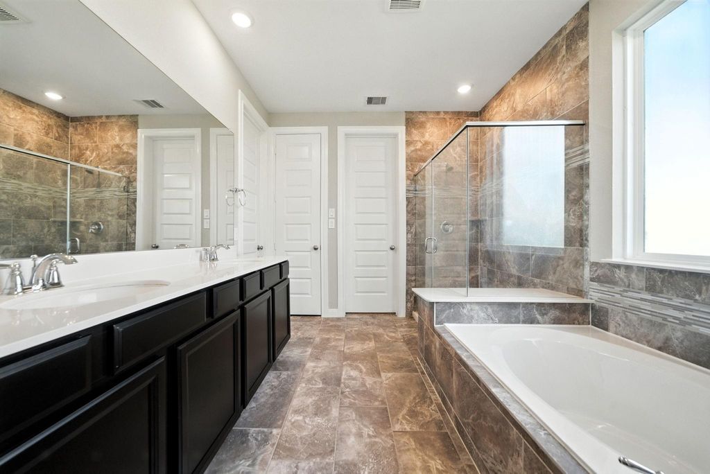 This modern bathroom features a sleek design with a double vanity, dark cabinetry, and a spacious countertop. It includes a glass-enclosed shower and a separate bathtub, both surrounded by elegant tile work. Natural light streams in through a large window, enhancing the room's bright and airy feel.